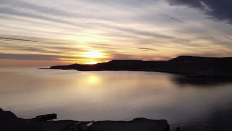 joven solitario sentado en una cornisa de montaña mirando al mar con una puesta de sol dorada - toma aérea con zoom