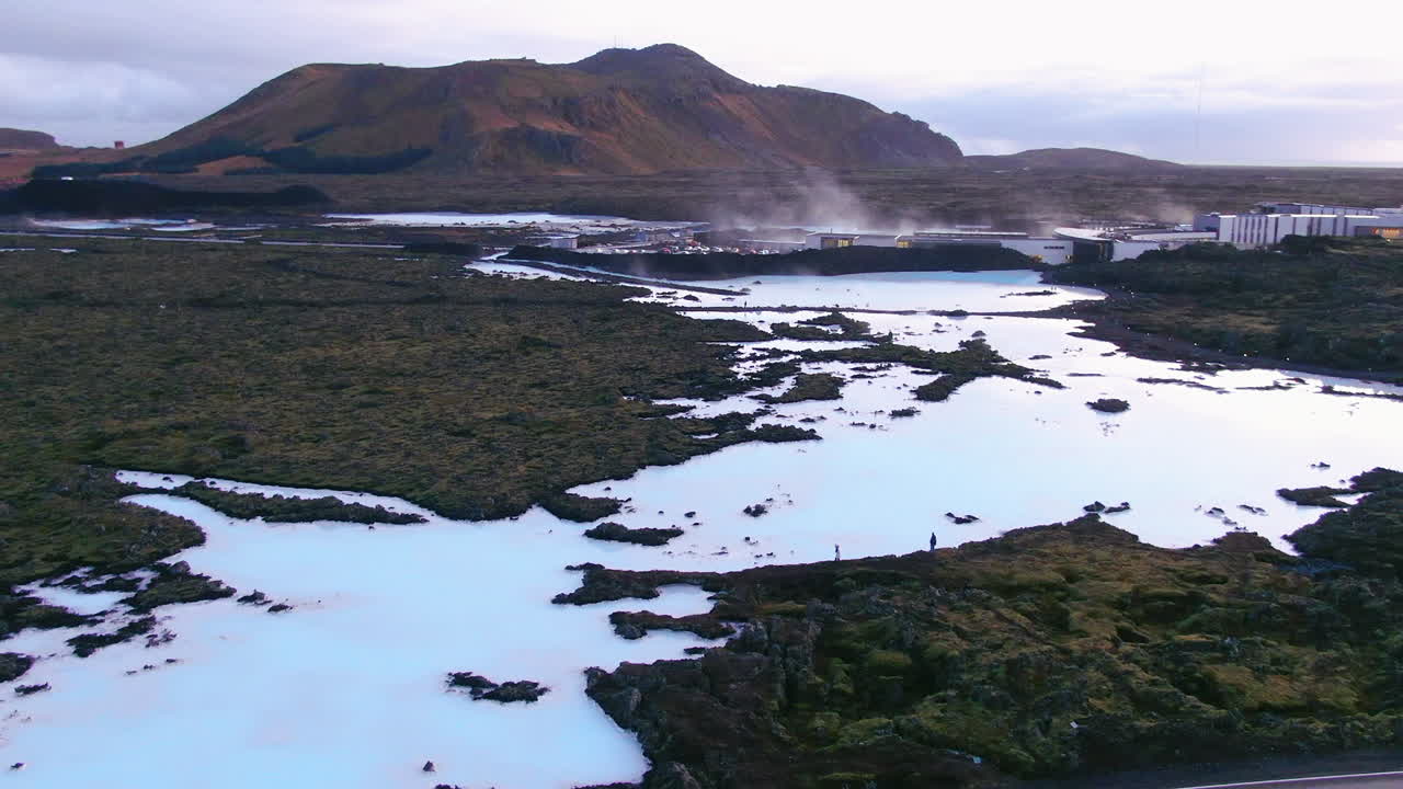 Blue lagoon partially frozen over with reflections of the sky, steam, roadway and mountains in the background in Iceland