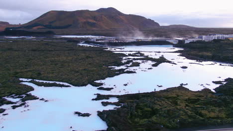 Blue-lagoon-partially-frozen-over-with-reflections-of-the-sky,-steam,-roadway-and-mountains-in-the-background-in-Iceland