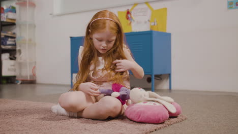 bambina rossa che raccoglie coniglio giocattolo mentre si siede sul pavimento in classe in una scuola montessori