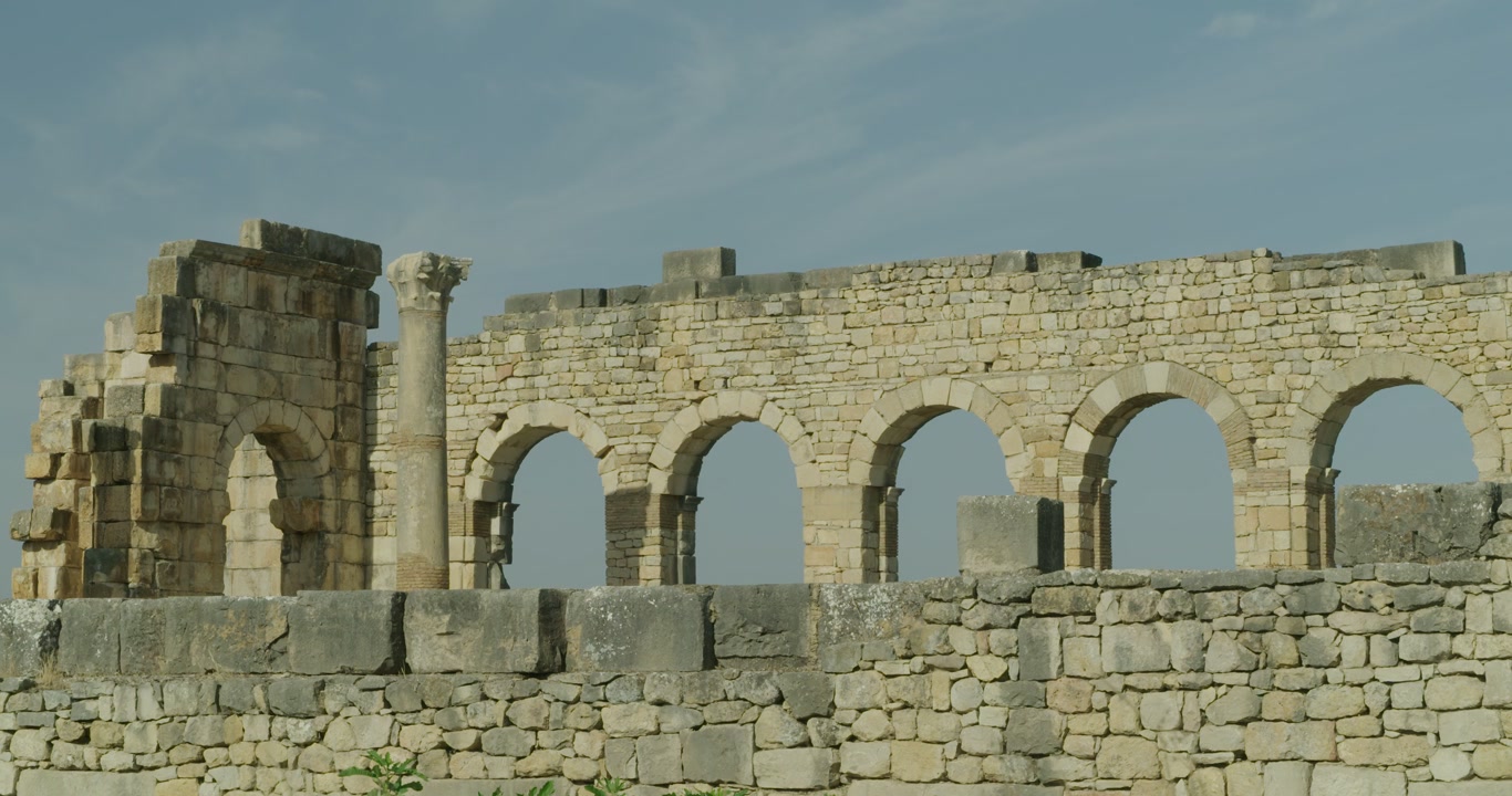 Vista cinematográfica amplia a nivel del suelo de la fachada con arcadas de la Basílica de Volubilis, destacando la monumental arquitectura cívica de uno de los sitios arqueológicos romanos mejor conservados de Marruecos.