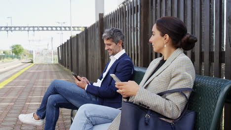 business commuters sitting on seat on railway platform with mobile phones