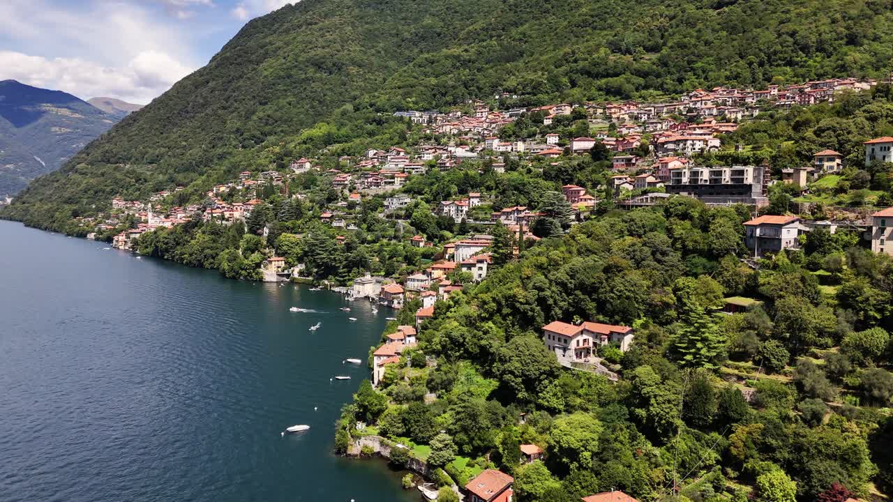 Nesso village on Lake Como's shoreline shows charming traditional Italian architecture climbing the lush green mountainside, with boats navigating the calm blue water, Lombardy, Italy