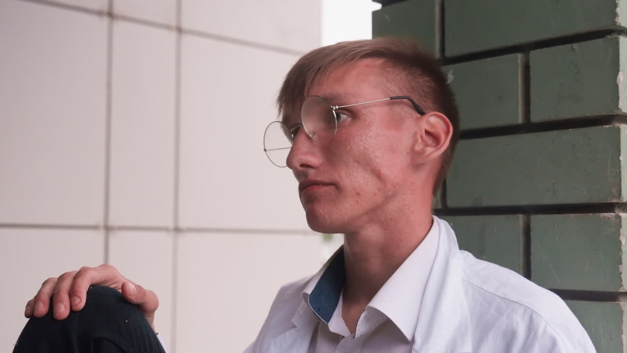 Biologist in white shirt wearing glasses eats pastry outdoors while seated near wall, reflecting casual moment of relaxation and hunger during break, with focus on thoughtful human expression