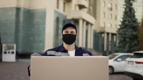 handsome delivery man in uniform, mask and gloves carrying big cardboard box parcel outdoor