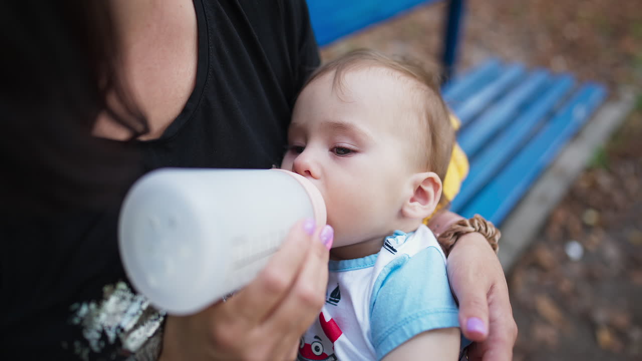 Alimentazione del bambino durante la passeggiata all'aperto. Dolce bambino che succhia il latte dal biberon e si strofina il piede. Primo piano. Sfondo sfocato