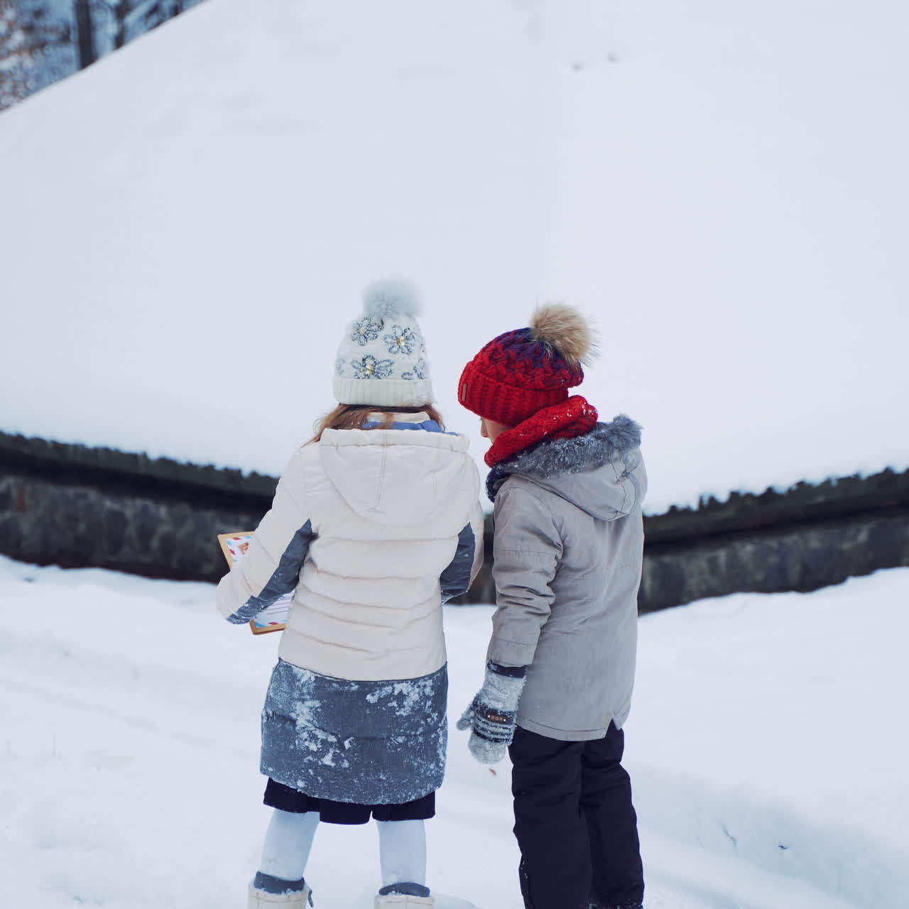Children stop near the snowy house and talk in winter. Back view of two little kids walking in beautiful white snow outdoors. Children in warm clothes look into Christmas card.