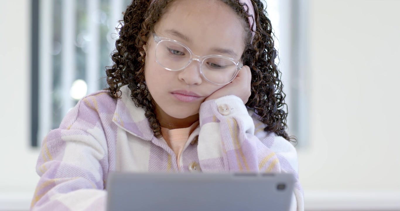 Bambina che guarda in alto, si sistema e appoggia la testa mentre lavora su un tablet a un tavolo con occhiali