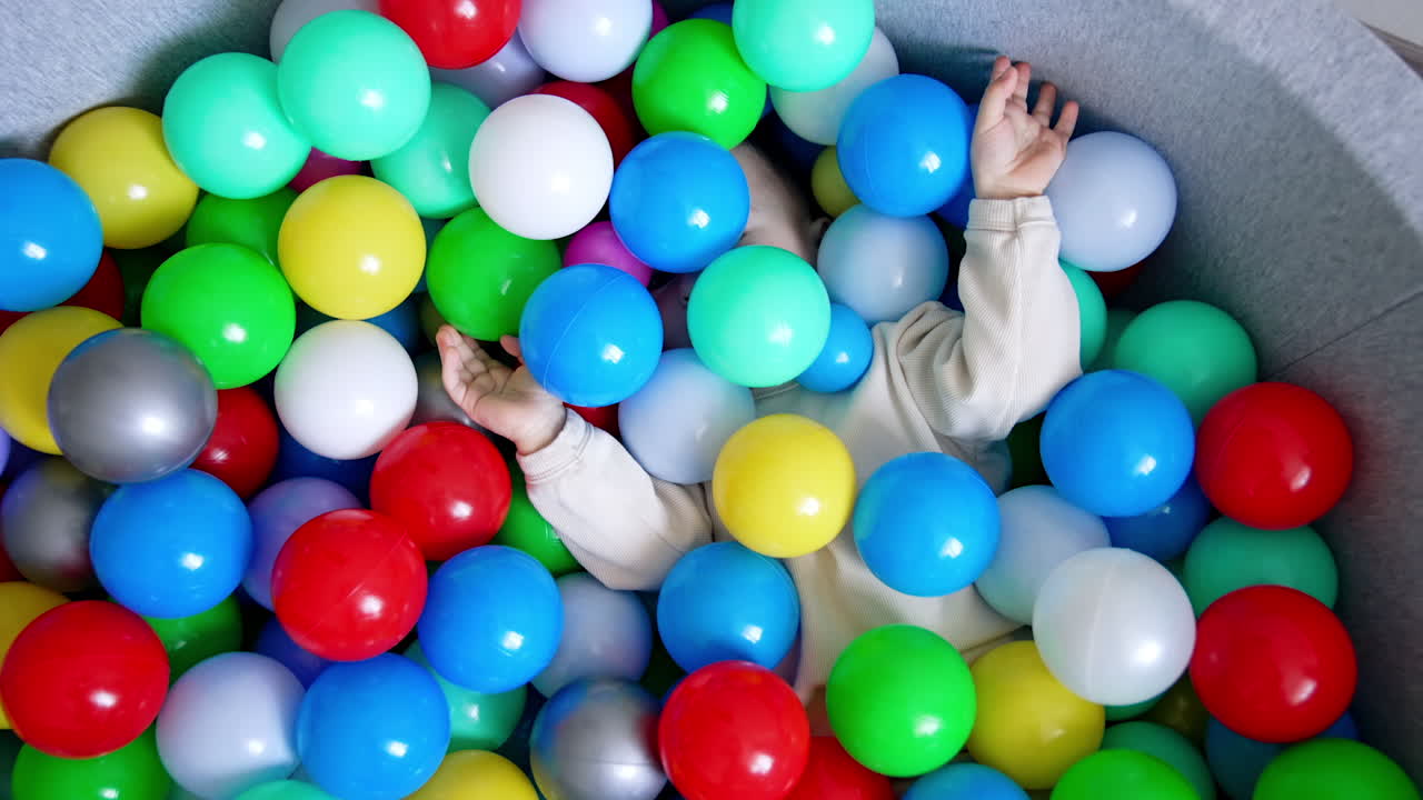 Baby boy lies hiding in the colorful balls in a dry pool. Toddler turns and sits in the basin taking one red ball in hands. Top view.