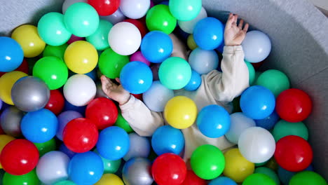 baby boy lies hiding in the colorful balls in a dry pool. toddler turns and sits in the basin taking one red ball in hands. top view.