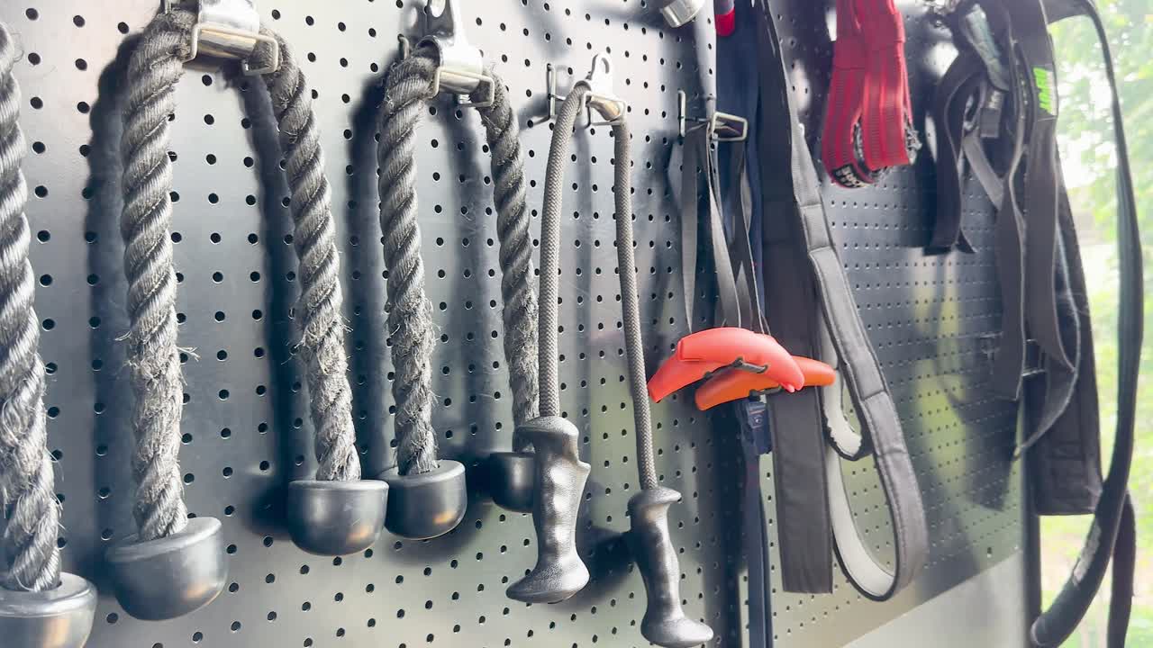 Various gym cable attachments and rope handles organized on pegboard, natural lighting, slow camera pan