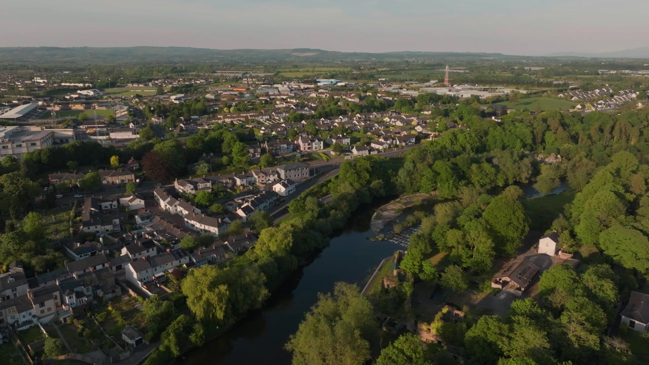 Una toma cinematográfica con dron que captura el exuberante paisaje verde, las viviendas residenciales y el apacible río Nore en Kilkenny, Irlanda, durante un cálido atardecer.
