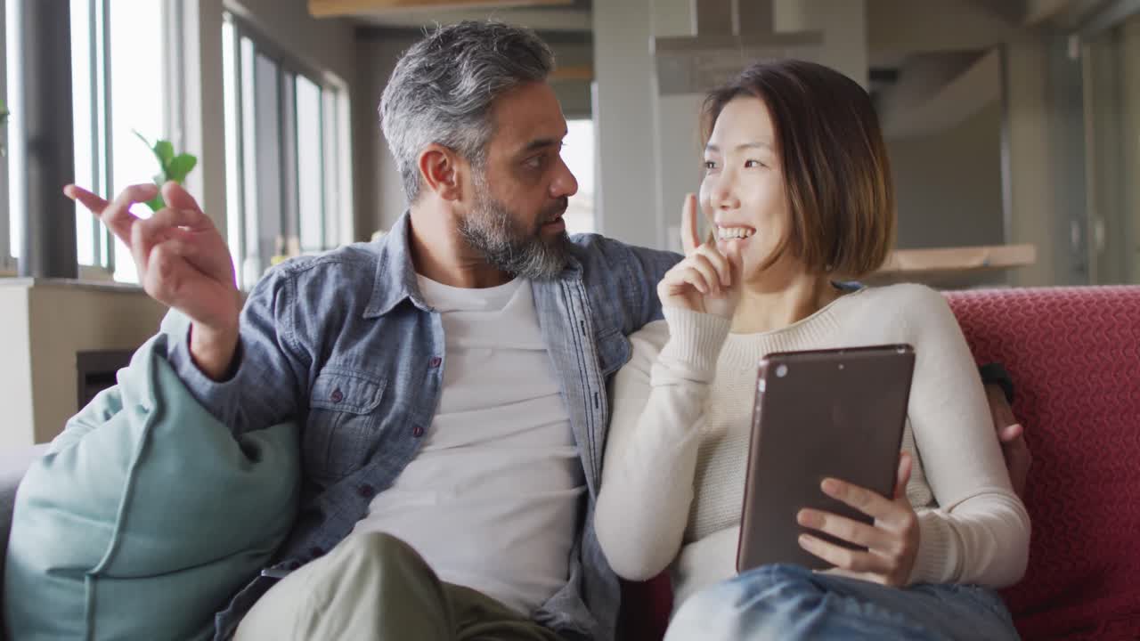 una pareja feliz y diversa sentada en la sala de estar con una tableta y hablando juntos