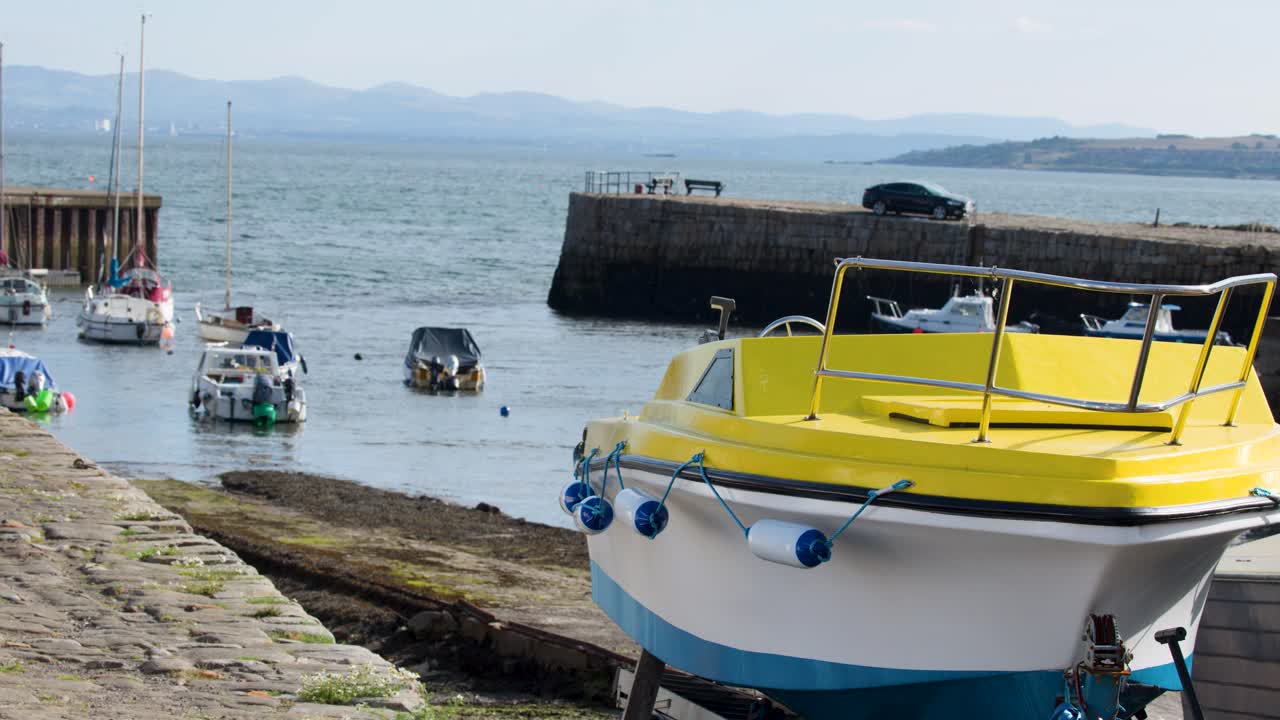 Static wide shot of colorful motorboats in calm harbor, bright daylight, stone slipway foreground