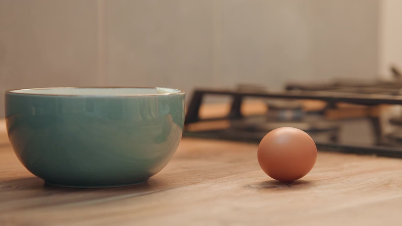 Una ciotola vuota attende accanto a un uovo marrone, ciotola di ceramica su un bancone di legno, una mano si estende nell'inquadratura, fornello a gas sfocato sullo sfondo, luce naturale calda, allestimento tranquillo del piano di lavoro per una ricetta.