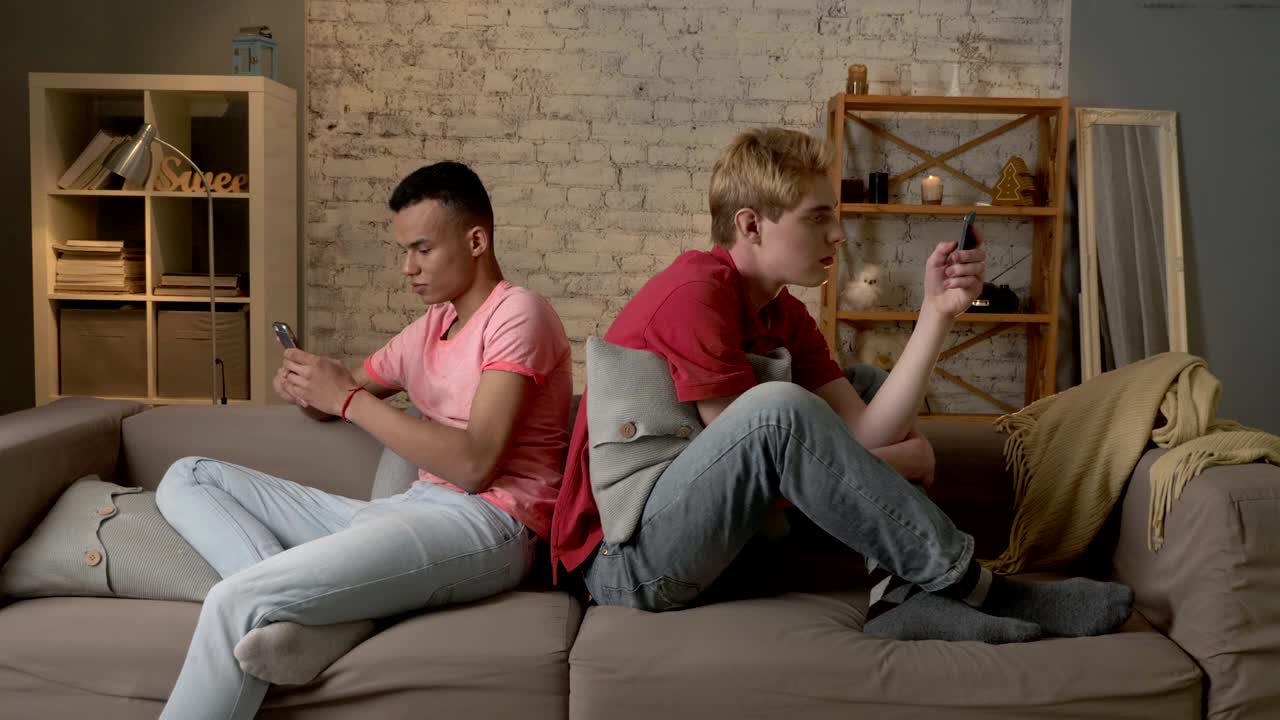 Two young mans are sitting on the couch and using a smartphones, gays, the problem of society, a new generation, lgbt lovers, homo, homosexuality concept 60 fps
