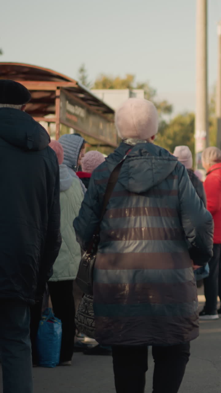 Caucasian seniors waiting at bus stop, crisp autumn morning, lined under shelter wearing warm coats and beanies, carrying bags and backpacks, soft sunlight on pavement, quiet suburban street, patient