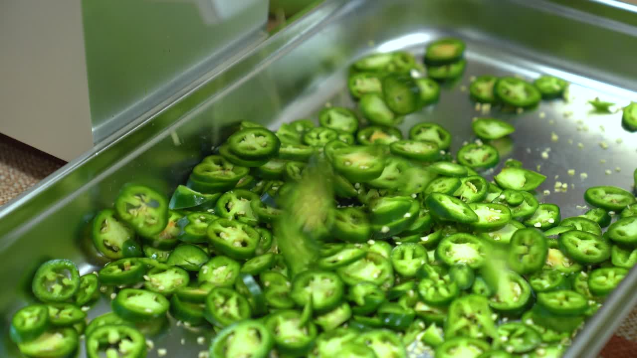 Jalapeños fatiados caindo em uma bandeja de um fatiador automático em cozinha comercial, fatiando pimentas Jalapeño verdes frescas para preparação de comida picante.