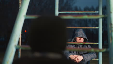 close-up of two individuals resting on an iron bar, with one in a hooded jacket looking away as light reflects off him, the background features a blurred building and a moving car