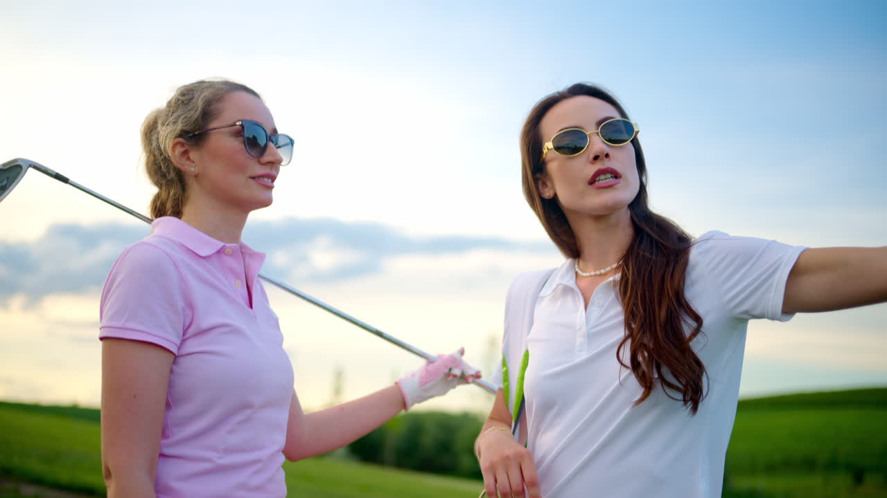 Two women dressed in white and pink clothes, holding golf clubs and talking on the course