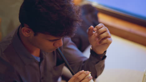 young man in buttoned shirt sits at wooden table, thoughtfully sipping drink through black straw while looking down, suggesting moment of quiet reflection, concentration