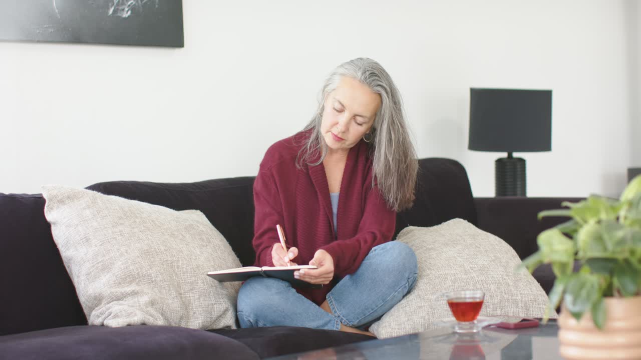 Donna anziana che tiene una penna, abbassa lo sguardo e scrive su un quaderno in soggiorno con una tazza di tè.