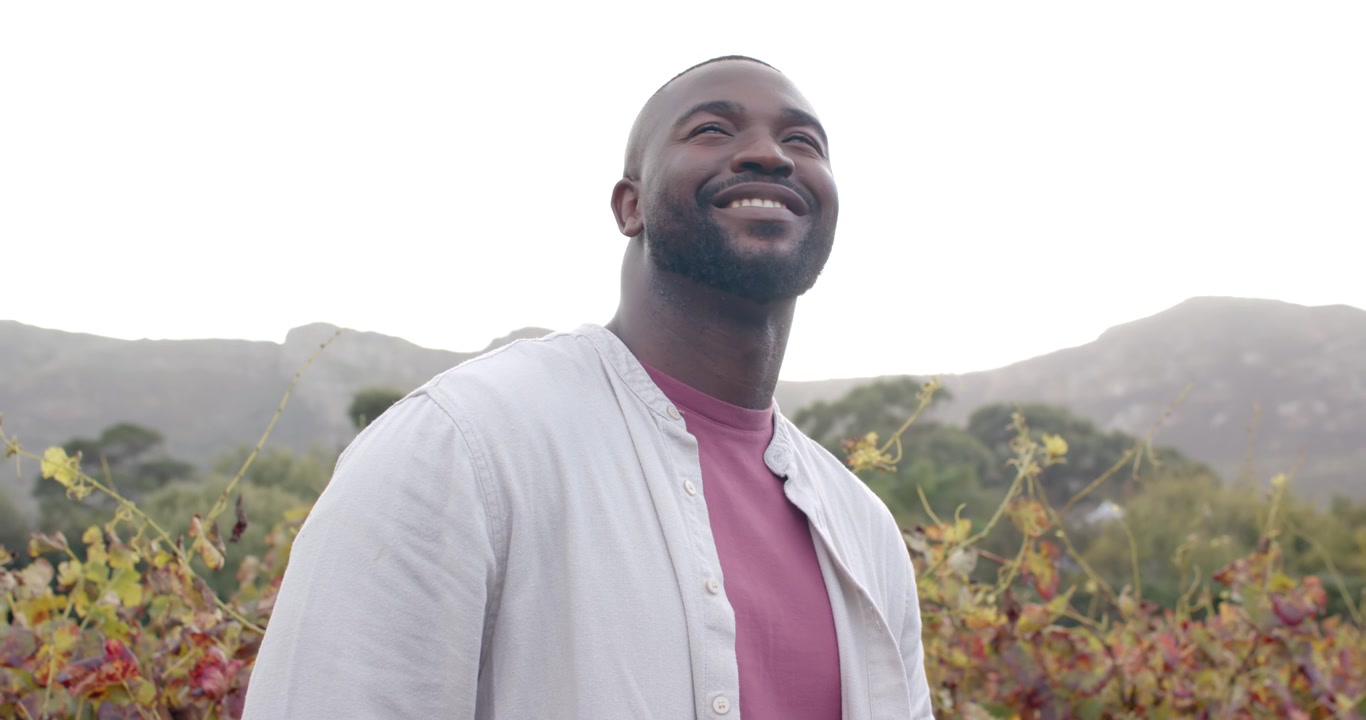 Adult African man standing in vineyard, looking upward at bright sky and smiling, wearing pink tee
