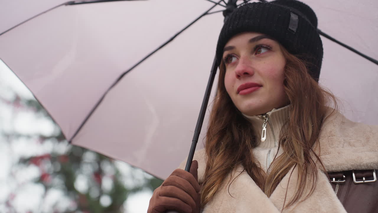 Primo piano di una ragazza allegra che cammina tenendo un ombrello, indossa un berretto di maglia e una giacca shearling marrone, sorride sotto una leggera nevicata con sfondo sfocato di alberi e ambiente urbano