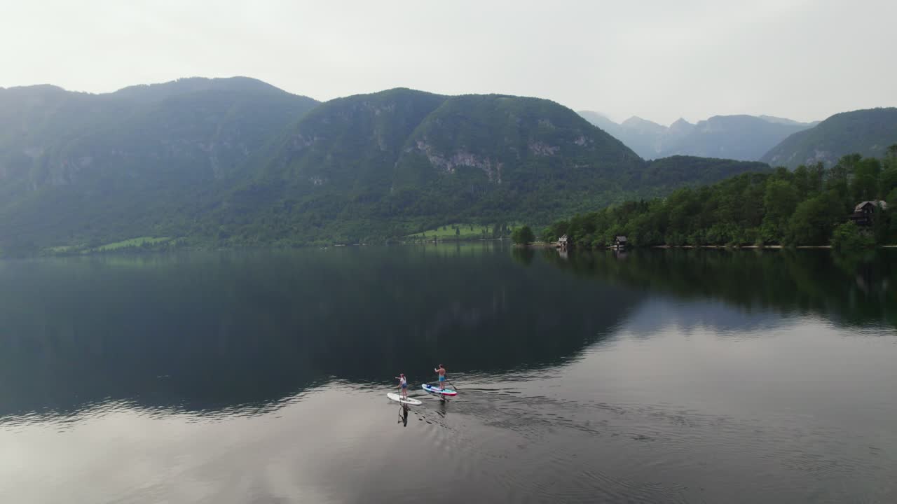 Ujęcie z drona nad jeziorem Bohinj w Słowenii, gdy para uprawia stand up paddling z górami w tle o zachodzie słońca