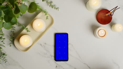 overhead view looking down on still life of blue screen mobile phone lit candles and incense stick with green plant and towels as part of spa day decor 2