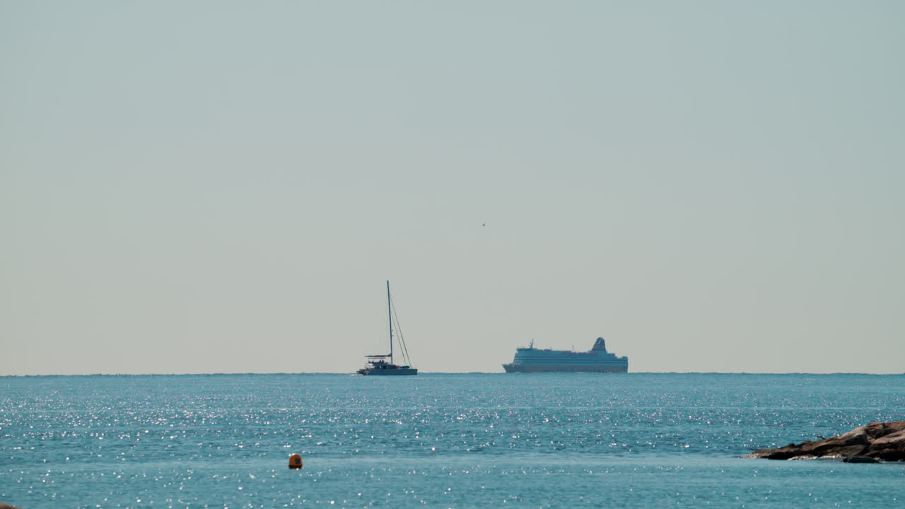 A cruise ship and a sailing boat move slowly on a shimmering blue sea under clear sunlight