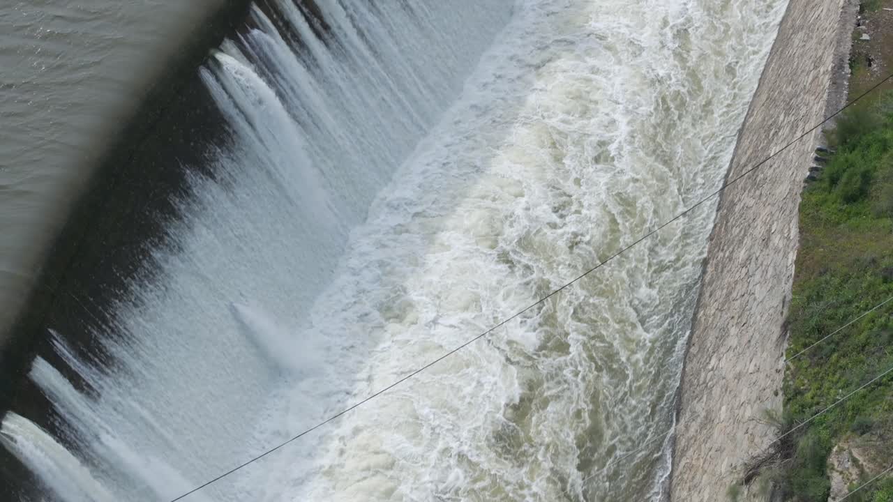 70mm drone footage of a dam spillway with a bridge in the foreground. Water cascades with great force into a wide channel. The drone flies opposite the flow, revealing a spectacular scene.