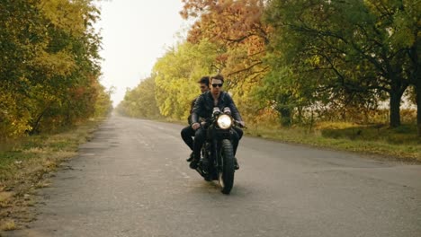 Handsome-man-in-sunglasses-sitting-with-his-girlfriend-behind-the-wheel-of-a-motorcycle-and-riding-on-the-asphalt-road-in-forest