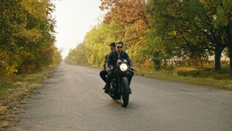 handsome man in sunglasses sitting with his girlfriend behind the wheel of a motorcycle and riding on the asphalt road in forest