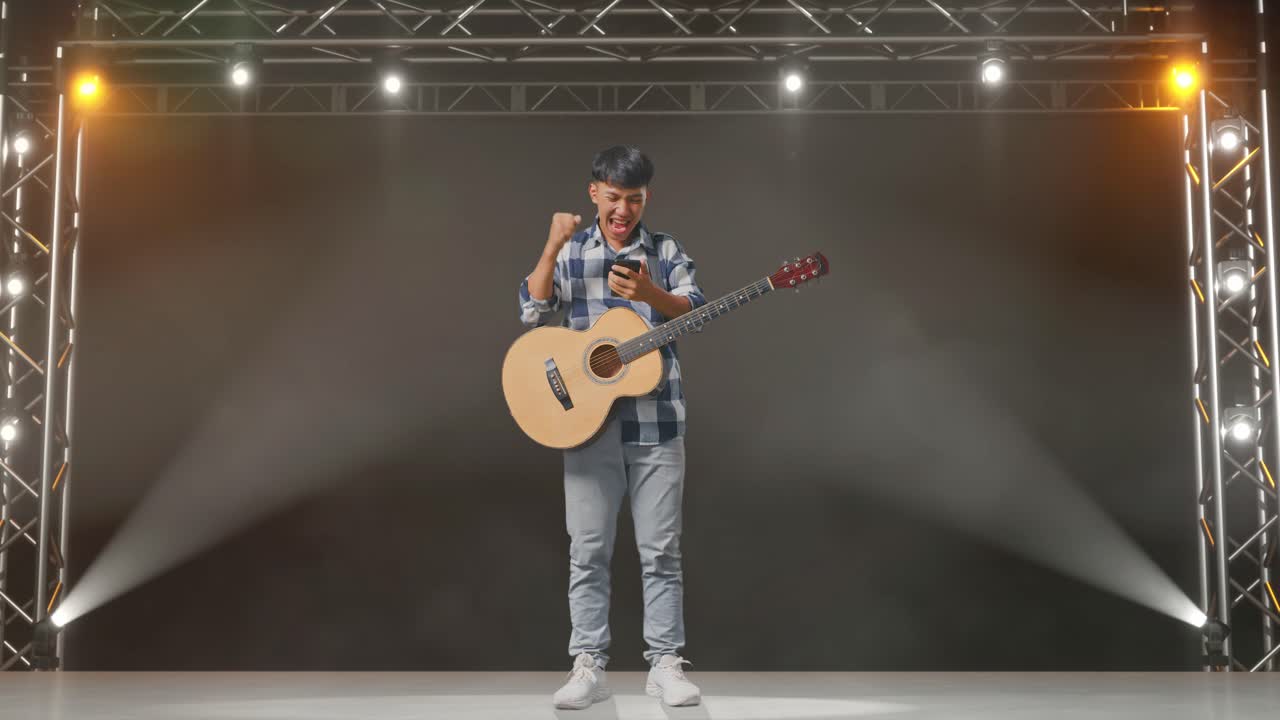 cuerpo lleno de adolescente asiático con guitarra usando teléfono inteligente y celebrando el éxito mientras está de pie en el escenario