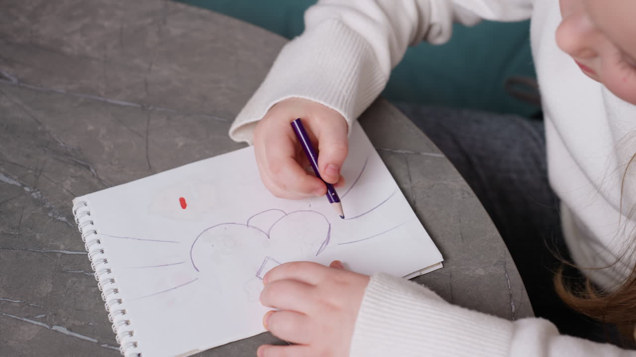 Close up of young girl drawing with purple pencil on white notebook at table, small hands holding sketchbook with child artwork, creative activity, childhood imagination, artistic expression