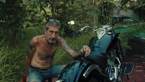 man with tattoos sitting by his motorcycle in a garden