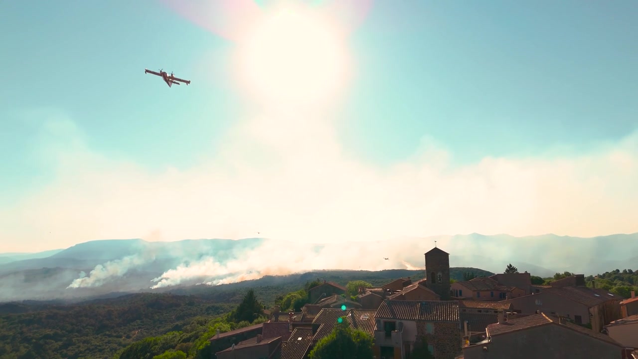 Dynamisk droneopptak fra lav vinkel som fanger et brannslukkingsfly av typen Canadair som flyr rett over hodet gjennom solens linseflare, med en massiv skogbrann og røyk som dekker åsene bak en historisk.