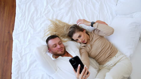 caucasian couple lies on the white bed sheets. beautiful woman taking selfie on her smartphone. top view.