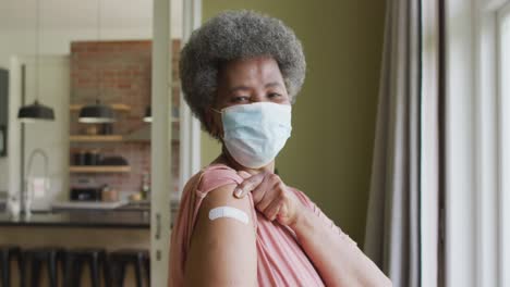 happy african american senior woman in face mask showing plaster on arm after covid vaccination