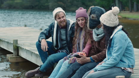 friends-using-smartphone-browsing-photos-together-sharing-memories-having-fun-sitting-on-jetty-by-lake-chatting-hanging-out-on-cloudy-winter-day-wearing-warm-clothes