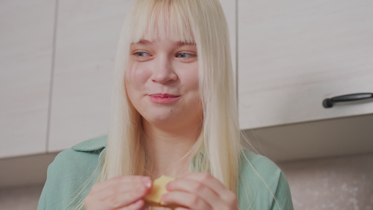 close up of cheerful blonde girl smiling and taking bite of sandwich while standing in bright modern kitchen with white cabinets behind her, hands holding bread, relaxed morning mealtime atmosphere