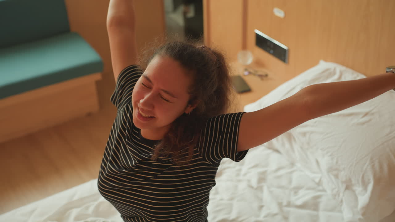 Una donna di colore si stira nel letto d'hotel, sbadigliando e sorridendo mentre alza le braccia verso il soffitto. La calda luce della finestra illumina il top a righe e il pigiama accogliente. Una viaggiatrice solitaria inizia la giornata con un'energia positiva.
