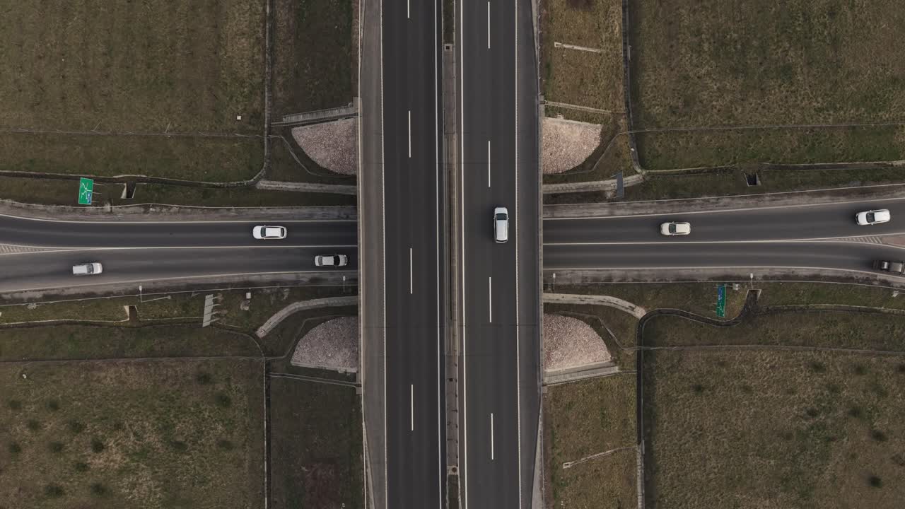 A top-down aerial view of a highway intersection with an overpass crossing above a main road. Several cars travel in both directions, surrounded by dry grassland and structured road layouts