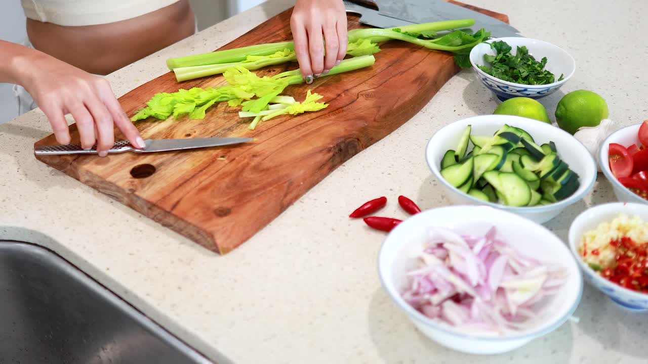 Hands chopping celery on a wooden board surrounded by fresh vegetables in a well-lit kitchen setting