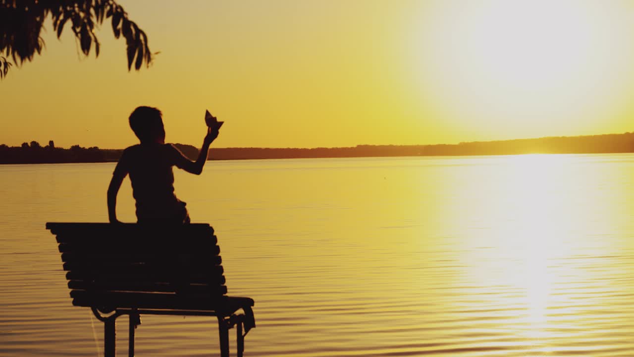 a little boy is sitting on the bench and playing with a paper boat on the background of evening landscape by the lake in the summer. Sunset
