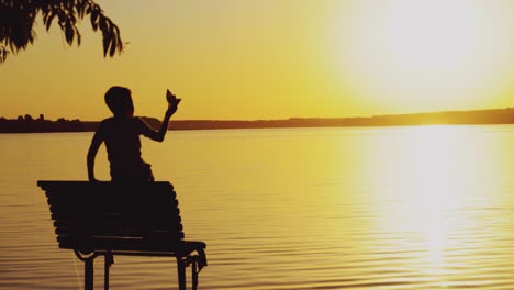 a little boy is sitting on the bench and playing with a paper boat on the background of evening landscape by the lake in the summer. sunset