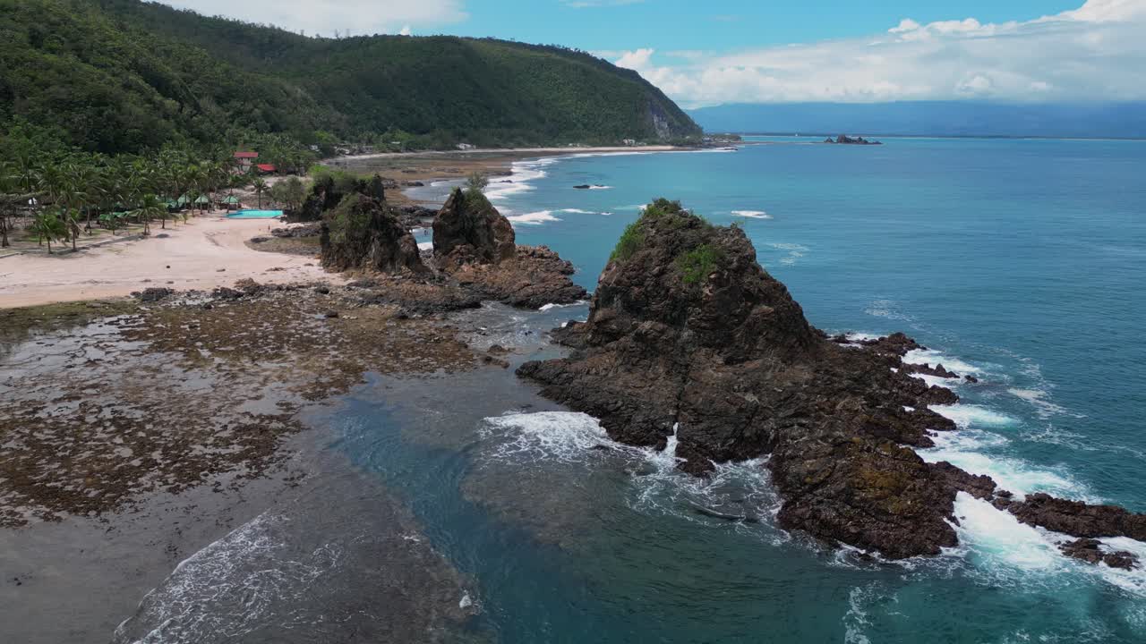 A serene drone shot reveals a small rocky mountain nestled near the shore, its jagged peaks contrasting beautifully with the smooth, sandy beach below.