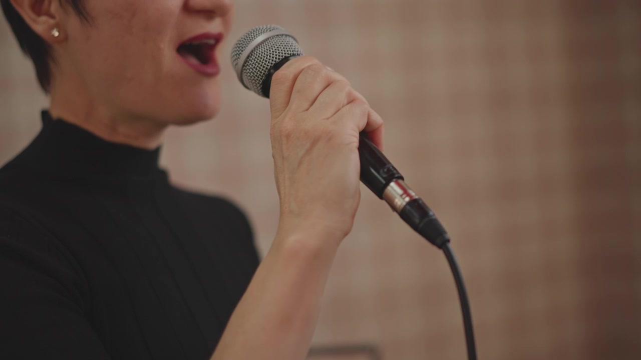 Pratica vocale solista in uno studio accogliente, riscaldamento guidato dall'insegnante con microfono a mano, sfondo a quadri, fraseggio espressivo, coaching sulla postura, allenamento della fiducia e prova di performance