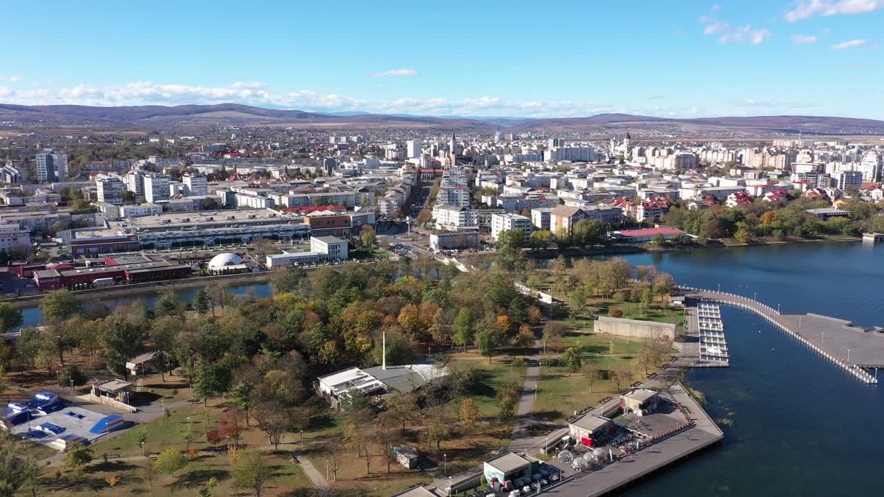 Una ripresa aerea lenta con drone mostra un vasto paesaggio urbano sulla riva di un grande lago blu. Bacău. Alberi autunnali punteggiano il parco sul lungomare sottostante, con montagne all'orizzonte.